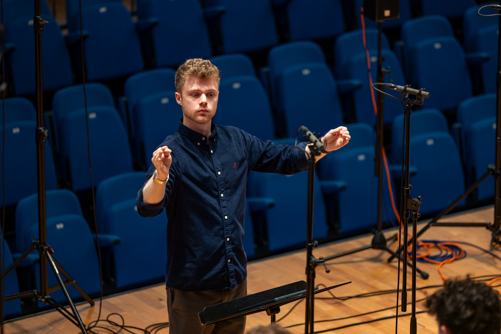 Adam Whitmore conducting with both arms raised in concert hall with blue seating